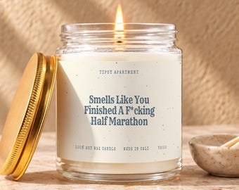 Könnte beinhalten: Eine brennende Sojawachskerze in einem klaren Glasgefäß mit goldenem Metalldeckel. Das Etikett trägt den Text: Tipsy Apartment, Smells Like You Finished A F*cking Half Marathon, 100% Soy Wax Candle, Made In Cali, T200. Die Kerze hat einen Docht.