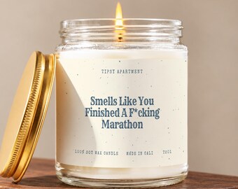 May include: A clear glass jar candle with a gold metal lid and a lit wick. The cream-colored label features the text Tipsy Apartment, Smells Like You Finished A F*cking Marathon, 100% Soy Wax Candle, Made In Cali, and the code T201 in dark blue.