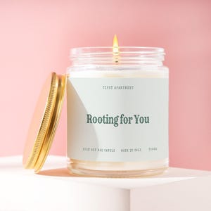 May include: A clear glass jar candle with a lit wick and a gold-colored metal lid placed to the side. The label is light green with dark green text reading Tipsy Apartment, Rooting for You, 100% soy wax candle, made in Cali, and T5662.