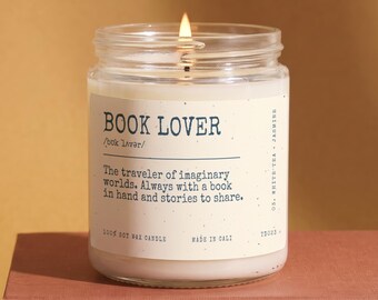 May include: A clear glass jar candle with a lit wick and white wax. The label features the text BOOK LOVER, a phonetic pronunciation, and the definition: The traveler of imaginary worlds. Always with a book in hand and stories to share. 100% soy wax.