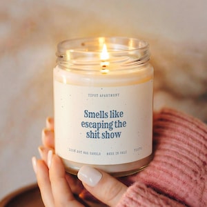 Pode incluir: Uma vela num frasco de vidro transparente com um pavio aceso e um rótulo cor de creme. O rótulo apresenta o texto "TIPSY APARTMENT", "Smells like escaping the shit show", "100% SOY WAX CANDLE", "MADE IN CALI" e "71270" em letras azul-escuras.