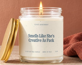 May include: A lit soy wax candle in a clear glass jar with a gold metal lid. The cream-colored label features the text "TIPSY APARTMENT", "Smells Like She's Creative As Fuck", "100% SOY WAX CANDLE", "MADE IN CALI", and the code "T165" in dark blue.