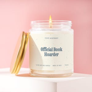 May include: A clear glass jar candle with a gold lid and a cream-colored label featuring the text "TIPSY APARTMENT", "Official Book Hoarder", "100% SOY WAX CANDLE", "MADE IN CALI", and "T1421". The candle has a lit wick and white wax inside.