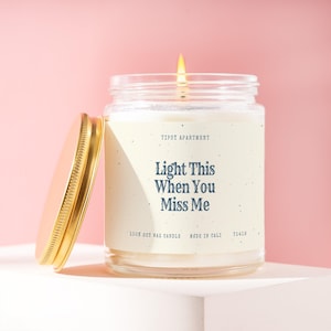 May include: A clear glass jar candle with a gold lid and a white label featuring the text "Tipsy Apartment", "Light This When You Miss Me", "100% soy wax candle", "made in Cali", and "T1416". The candle has a lit wick with a visible flame.
