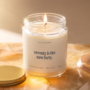 May include: A clear glass jar candle with a lit wick and a cream-colored label. The label features the text "TIPSY APARTMENT" at the top, "seventy is the new forty." in the center, and "100% SOY WAX CANDLE MADE IN CALI T1246" at the bottom.
