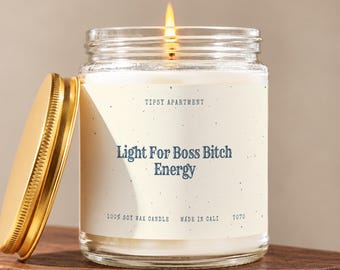 Puede incluir: Una vela en frasco de vidrio transparente con una mecha encendida y una tapa de metal dorado. La etiqueta es de color crema con texto azul oscuro que dice: Tipsy Apartment, Light For Boss Bitch Energy, 100% Soy Wax Candle, Made In Cali y T070.