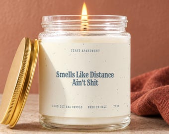 May include: A lit soy wax candle in a clear glass jar with a gold-colored metal lid. The label features the text Tipsy Apartment, Smells Like Distance Ain't Shit, 100% Soy Wax Candle, Made in Cali, and T133 printed in dark blue on a cream background.