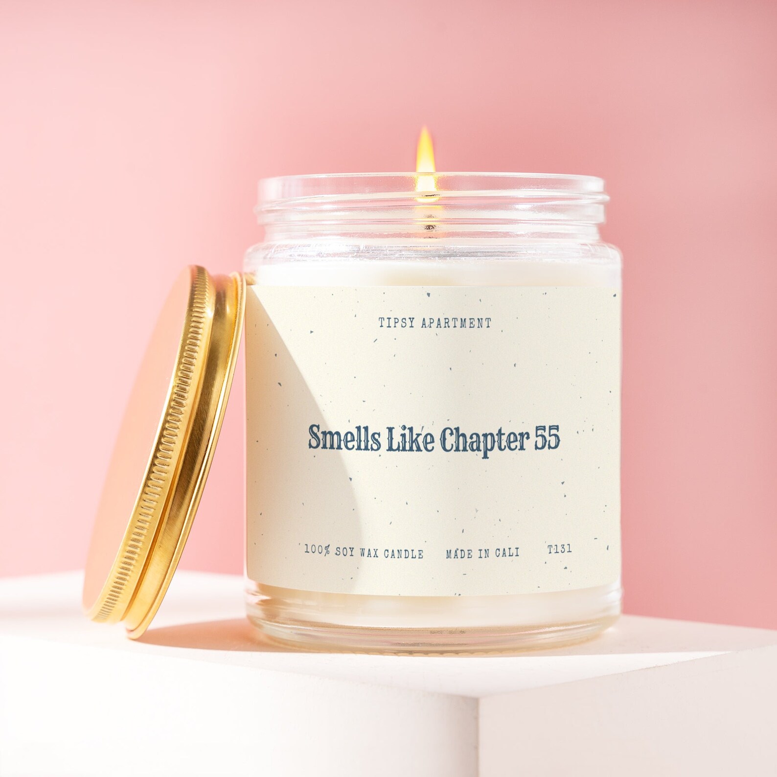 An open candle on a white surface against a pink background. The label says "Smells Like Chapter 55."