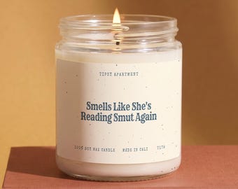 May include: A clear glass jar candle with a white wax fill and a lit wick. The label is cream-colored with dark blue text reading Tipsy Apartment, Smells Like She's Reading Smut Again, 100% Soy Wax Candle, Made in Cali, and the code T179.
