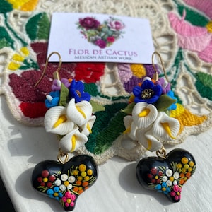 Op de afbeelding: Een paar oorhangers met een cluster van witte, blauwe en gele polymer clay bloemen met een zwarte hartvormige hanger. De hartvormige hanger is beschilderd met kleurrijke stippen en swirls. De oorhangers zijn handgemaakt en hebben goudkleurige haakjes.