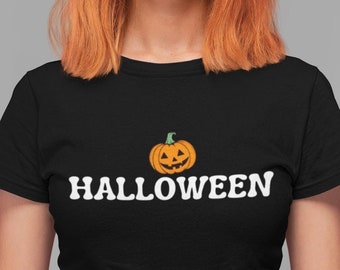 May include: A black short-sleeved t-shirt featuring a graphic of an orange jack-o'-lantern pumpkin centered above the white text HALLOWEEN in a bold, rounded font. The shirt has a crew neckline and is worn tucked into dark denim pants.