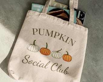 May include: Beige tote bag with the text "PUMPKIN Social Club" and illustrations of pumpkins. The bag contains magazines. The pumpkins are white and orange with green stems. The bag is made of a light-colored fabric.