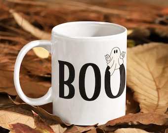 May include: A white ceramic mug featuring the black text BOO printed on the side. The letter O is replaced by a small white ghost illustration with black eyes and two hands making peace signs. The mug has a curved handle on the left side.