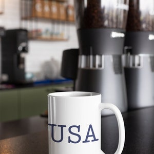 May include: White ceramic mug with the word "USA" in navy blue lettering. The mug is on a dark surface, with coffee grinders and coffee beans in the background. The scene is set in a coffee shop.
