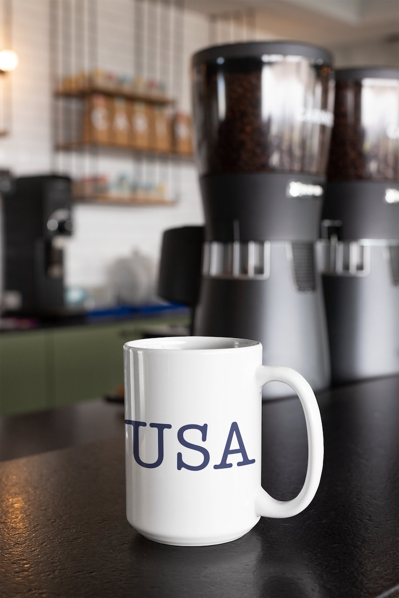 May include: White ceramic mug with the word "USA" in navy blue lettering. The mug is on a dark surface, with coffee grinders and coffee beans in the background. The scene is set in a coffee shop.