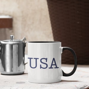 May include: White ceramic mug with the word "USA" in blue lettering. The mug has a black interior and a black handle. A stainless steel teapot is to the left of the mug.