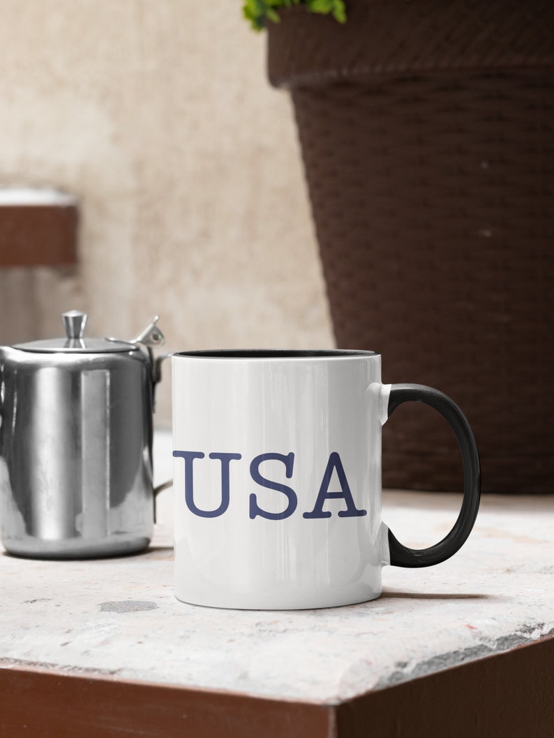 May include: White ceramic mug with the word "USA" in blue lettering. The mug has a black interior and a black handle. A stainless steel teapot is to the left of the mug.