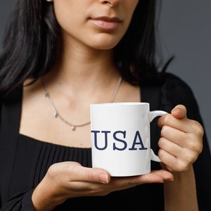 May include: White ceramic mug with the word "USA" printed in navy blue. The mug has a standard handle and is held by a person. The background is a solid gray.
