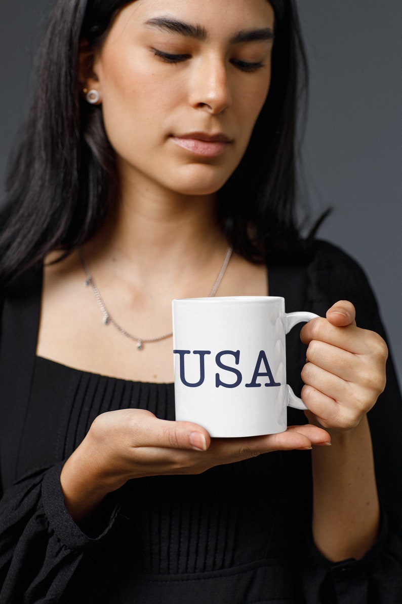 May include: White ceramic mug with the word "USA" printed in navy blue. The mug has a standard handle and is held by a person. The background is a solid gray.