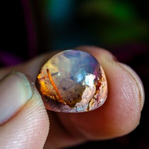 May include: A small, polished cabochon gemstone featuring a translucent, dome-shaped top with iridescent flashes of blue and white, transitioning into a textured, earthy base with warm orange and brown inclusions visible within the stone.