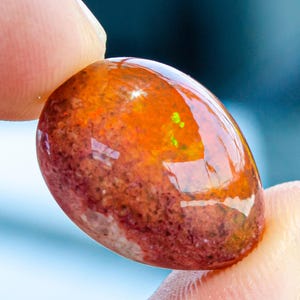 Puede incluir: Una gema pulida, de forma ovalada, sostenida entre los dedos. La piedra presenta una mezcla de colores, incluyendo naranja, marrón y toques de verde. La superficie es brillante, reflejando la luz. La gema parece ser un tipo de ópalo.
