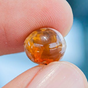 Puede incluir: Una pequeña piedra preciosa translúcida y redondeada con un tono ámbar y naranja intenso. La piedra presenta reflejos de luz internos y una superficie lisa y pulida, mostrando una paleta de colores cálidos y ardientes en su forma circular.