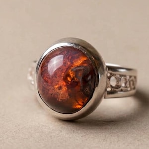 Puede incluir: Anillo de plata con una piedra grande, ovalada, de color marrón rojizo. La piedra tiene un aspecto veteado con zonas más claras y oscuras. La banda del anillo tiene un patrón decorativo de pequeñas formas recortadas.