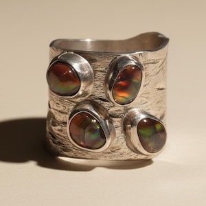 Puede incluir: Anillo ancho de plata con superficie texturizada, adornado con cinco piedras de ammolita ovaladas. Las piedras de ammolita exhiben un vibrante juego de colores, incluyendo tonos rojos, naranjas y verdes, engastadas en biseles individuales.