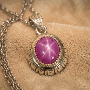 Puede incluir: Un collar de plata con un colgante con una gran gema rosa ovalada en forma de estrella. El colgante está engastado en un marco de plata decorativo con un diseño detallado. La cadena es de estilo eslabón clásico.