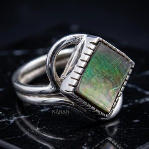 Puede incluir: Un anillo de plata con una incrustación cuadrada de concha de abulón iridiscente con textura agrietada. La banda de metal tiene un acabado pulido y un borde decorativo con muescas alrededor de la piedra. La palabra KANAN está grabada.