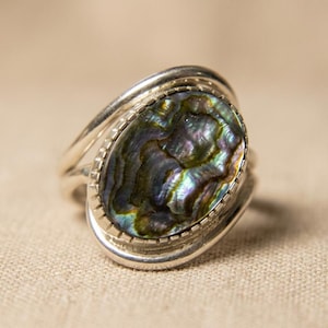 Puede incluir: Un anillo de plata con una concha de abulón ovalada. La concha iridiscente muestra tonos de verde, azul y morado. El anillo tiene un diseño de doble banda y un engaste biselado texturizado.
