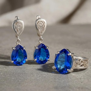 Puede incluir: Un conjunto de joyas de plata con un par de pendientes de lágrima y un anillo a juego. Cada pieza muestra una gran gema azul vibrante. Los engastes del anillo y los pendientes tienen detalles decorativos intrincados.