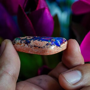 May include: A blue, green, and purple opal gemstone with a copper-colored base. The opal is a cabochon cut and is being held by a hand.