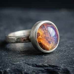 May include: A silver ring featuring a round, bezel-set cabochon stone with a mottled, translucent amber and orange interior. The metal band has a textured, matte finish, showcasing a rustic appearance with visible surface irregularities.