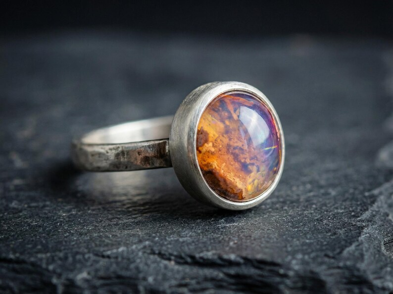 May include: A silver ring featuring a round, bezel-set cabochon stone with a mottled, translucent amber and orange interior. The metal band has a textured, matte finish, showcasing a rustic appearance with visible surface irregularities.