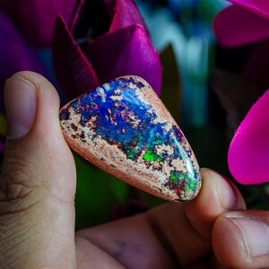 May include: A triangular shaped opal gemstone with a brown, white, blue, green, and purple iridescent sheen. The opal is being held by a hand.