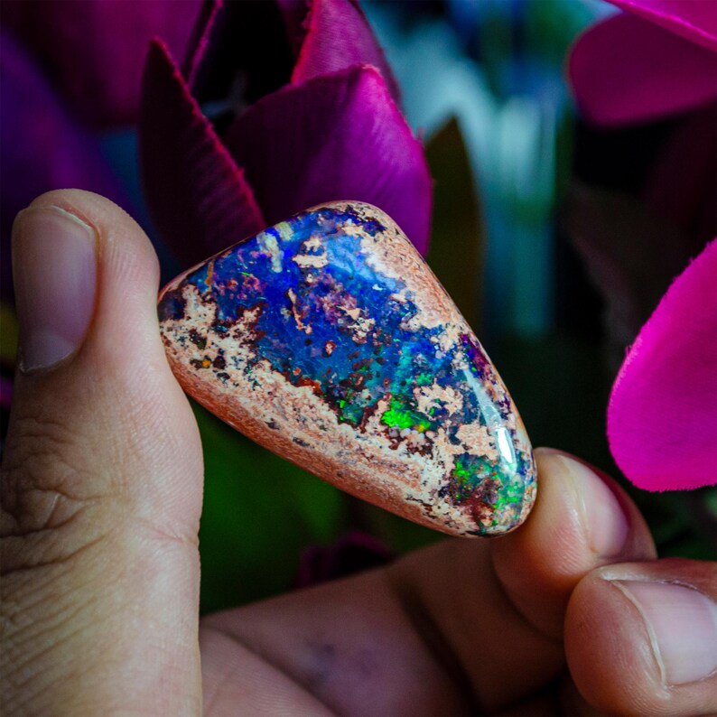 May include: A triangular shaped opal gemstone with a brown, white, blue, green, and purple iridescent sheen. The opal is being held by a hand.