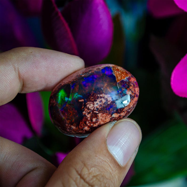 May include: A vibrant, iridescent opal cabochon with a mix of green, blue, and purple hues. The stone has a rough, natural texture and a dark brown base.