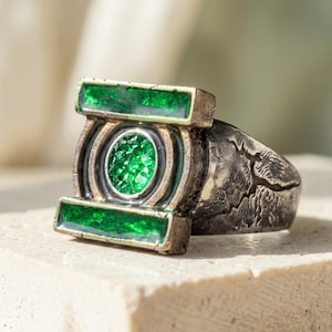 Puede incluir: Un anillo de tono plateado con una banda texturizada y desgastada, y un emblema rectangular central. El emblema muestra un centro cristalino verde circular flanqueado por dos incrustaciones rectangulares verdes, similar a una batería.
