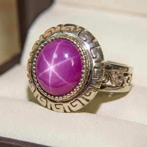 Puede incluir: Un anillo de plata con una gran gema de rubí estrella ovalada de color rosa púrpura. El anillo tiene un diseño ornamentado y grabado alrededor de la gema y en la banda. El anillo se muestra en una caja de joyería blanca.