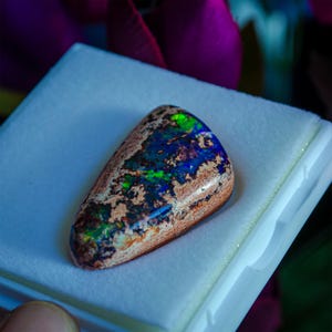 May include: A teardrop-shaped opal gemstone with a brown, white, and black matrix. The opal displays a vibrant blue, green, and purple iridescence.