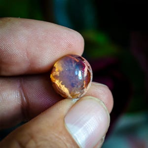 May include: A small, round, translucent gemstone with a smooth, polished surface. The stone displays a play of color with internal flashes of orange, yellow, and deep red hues against a cloudy, pale base, creating a vibrant, multi-colored effect.