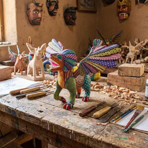 May include: A hand-carved wooden alebrije sculpture featuring a mythical creature with vibrant, multi-coloured geometric patterns, large wings, and a fierce expression, displayed on a workbench surrounded by carving tools and paint supplies.