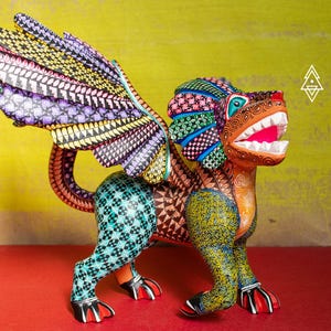 May include: A hand-painted wooden alebrije sculpture featuring a winged creature with a lion-like head, sharp white teeth, and a vibrant, multi-coloured body covered in intricate geometric patterns, including shades of yellow, teal, and purple.