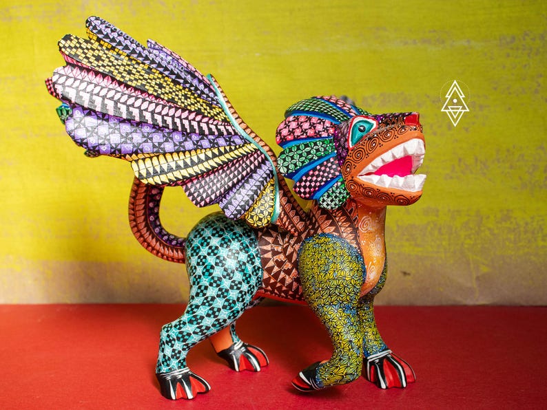May include: A hand-painted wooden alebrije sculpture featuring a winged creature with a lion-like head, sharp white teeth, and a vibrant, multi-coloured body covered in intricate geometric patterns, including shades of yellow, teal, and purple.