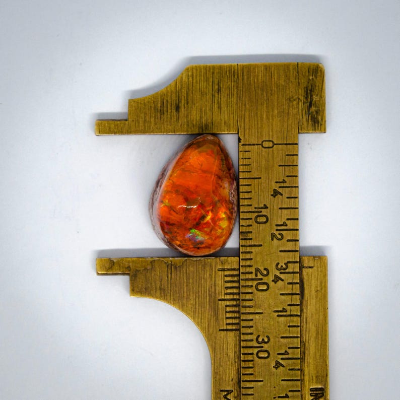 May include: A vibrant, teardrop-shaped gemstone, primarily orange with hints of red and iridescent flashes. The stone is measured with a brass ruler, appearing to be approximately 0.75 inches in length. The gemstone is set against a white background.