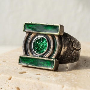 Puede incluir: Un anillo de tono plateado con un diseño rectangular, un elemento circular central con textura cristalina verde y dos incrustaciones rectangulares verdes horizontales, todo sobre una banda metálica oscura y desgastada.