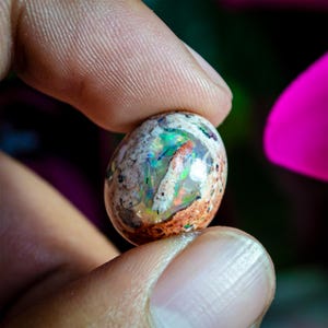 Puede incluir: Una piedra preciosa de ópalo boulder de forma ovalada y pulida, con una matriz de color marrón tierra texturizada y destellos vibrantes de juego de colores iridiscentes en verde, azul y rojo en el centro translúcido.