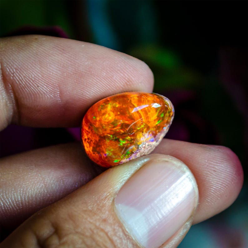 May include: A vibrant, polished opal gemstone, held between fingers. The opal displays a fiery orange hue with internal flashes of green and yellow, creating a captivating play of color. The stone has a smooth, rounded shape.