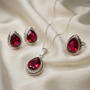 Puede incluir: Un conjunto de joyas que incluye un collar de plata con colgante, pendientes a juego y un anillo. Cada pieza presenta una piedra preciosa roja oscura en forma de lágrima con un borde plateado decorativo. El conjunto se exhibe sobre una tela blanca y sedosa.